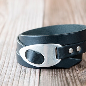 May include: A black leather wrap bracelet with a silver oval-shaped buckle. The bracelet is on a wooden surface.