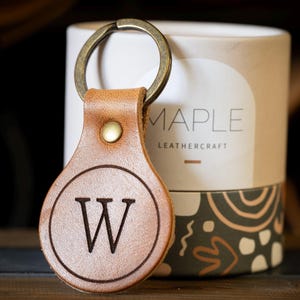 Op de afbeelding: Een beige leren sleutelhanger met een ronde hanger voorzien van een in reli&euml;f aangebrachte letter W in een cirkel. De sleutelhanger is bevestigd aan een bronskleurige metalen ring en een koperen klinknagel. Erachter staat een cilindrische houder met het opschrift MAPLE LEATHERCRAFT.