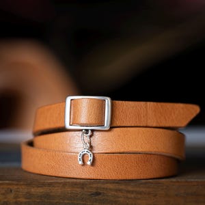 May include: A tan leather wrap bracelet featuring a rectangular silver-toned metal buckle and a small silver-toned horseshoe charm dangling from the centre. The smooth leather strap is coiled to create a layered appearance around the wrist.