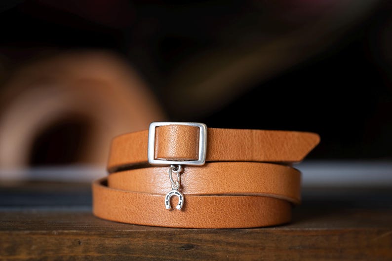 May include: A tan leather wrap bracelet featuring a rectangular silver-toned metal buckle and a small silver-toned horseshoe charm dangling from the centre. The smooth leather strap is coiled to create a layered appearance around the wrist.