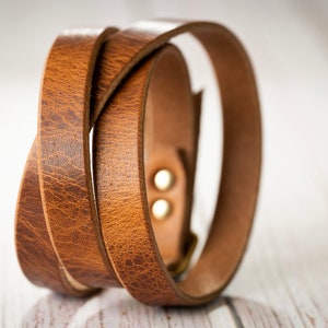 May include: A wrap-style bracelet crafted from textured brown leather. The band features a visible grain pattern and is secured with two metallic studs. The leather is looped to create a layered appearance, showcasing the natural tan edges and finish.