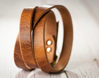 May include: A wrap-style bracelet crafted from textured brown leather. The band features a visible grain pattern and is secured with two metallic studs. The leather is looped to create a layered appearance, showcasing the natural tan edges and finish.