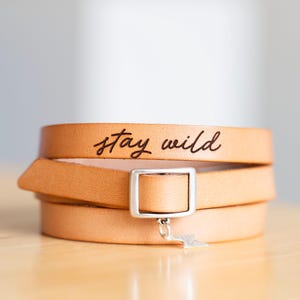 May include: A tan leather wrap bracelet featuring the text stay wild engraved in black script. The bracelet includes a rectangular silver-toned metal buckle and a small silver-toned lightning bolt charm hanging from the centre of the buckle.