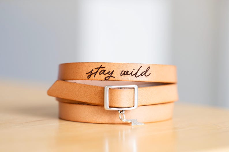 May include: A tan leather wrap bracelet featuring the text stay wild engraved in black script. The bracelet includes a rectangular silver-toned metal buckle and a small silver-toned lightning bolt charm hanging from the centre of the buckle.