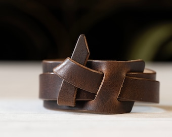 May include: A brown leather bracelet with a braided design. The bracelet is made of two strands of leather that are woven together to create a unique and stylish look.