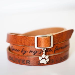 May include: A brown leather wrap bracelet with a silver buckle and a paw print charm. The bracelet is engraved with the words "Once by my side, forever" and the name "Oliver" and dates.