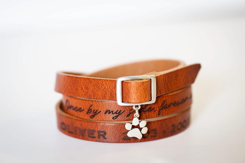 Close-up of a handcrafted triple-wrap leather bracelet in warm brown, featuring the engraved quote "once by my side, forever in my heart," with a sterling silver paw charm hanging from the buckle. Personalized with pet's name and dates for a custom memorial gift. Adjustable 24-inch strap or custom sizing. Handmade by Maple Leathercraft, perfect for pet remembrance and sympathy gifts.