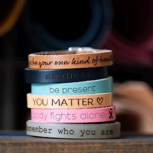May include: A stack of leather bracelets in various colors, each engraved with an inspirational message. The bracelets include the phrases "Be your own kind of beauty," "Be the light," and "You Matter." The bracelets are stacked on a wooden surface.