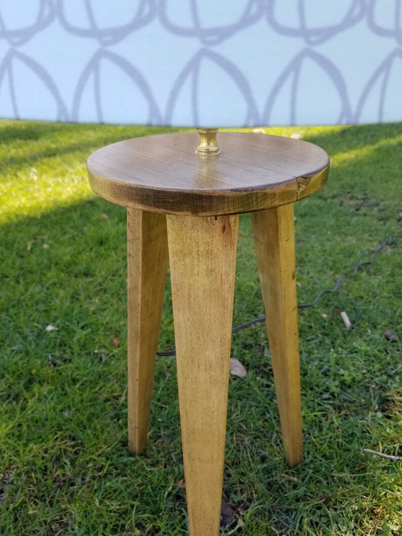 May include: A wooden stool with three legs and a round top. The stool is stained a light brown color and has a brass fitting on top.