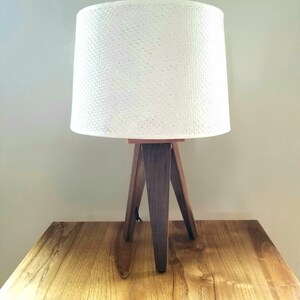 May include: A white lampshade with a woven texture sits atop a wooden tripod lamp base. The lamp is on a wooden table.