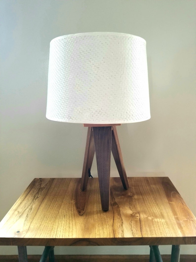 May include: A white lampshade with a woven texture sits atop a wooden tripod lamp base. The lamp is on a wooden table.