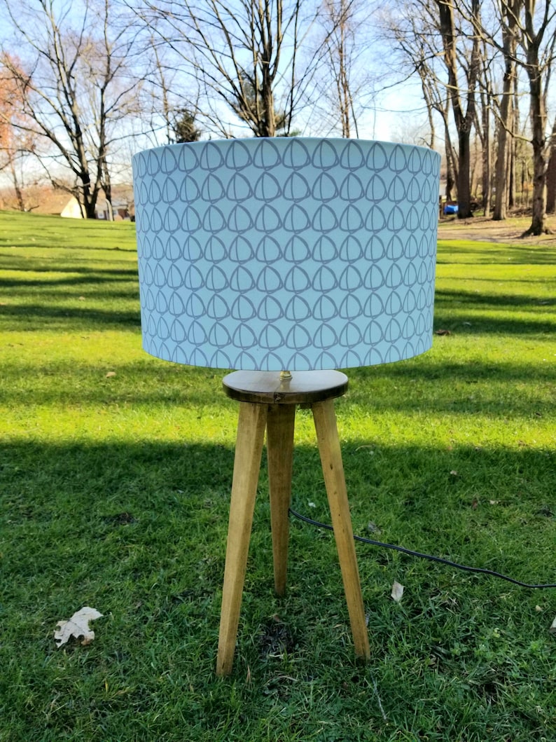 May include: A tripod lamp with a light blue lampshade featuring a repeating pattern of gray circles. The lampshade is made of fabric and has a white background. The lamp is on a wooden base and has a black cord.