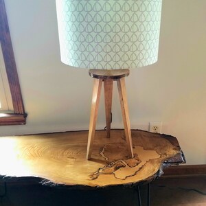 May include: A wooden tripod lamp with a light green lampshade featuring a gray geometric pattern. The lamp is sitting on a large, round wooden table with a natural wood grain.