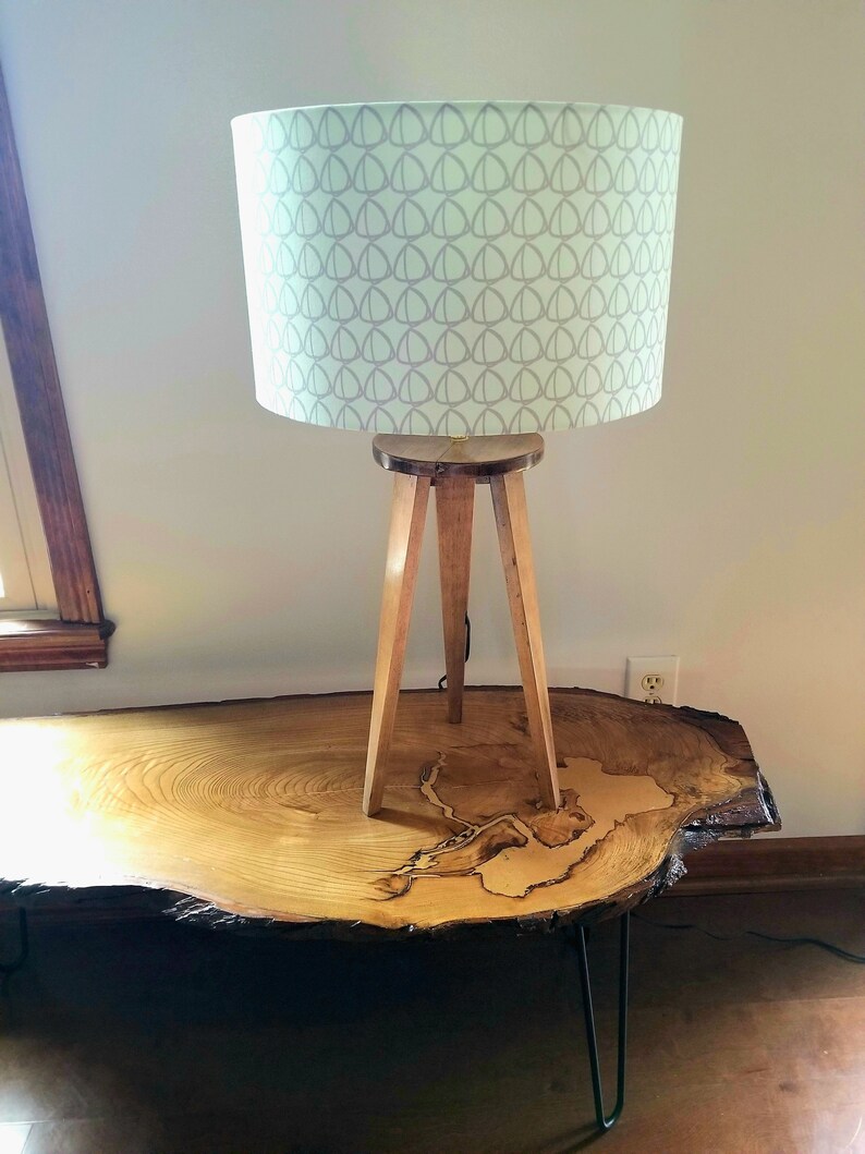 May include: A wooden tripod lamp with a light green lampshade featuring a gray geometric pattern. The lamp is sitting on a large, round wooden table with a natural wood grain.