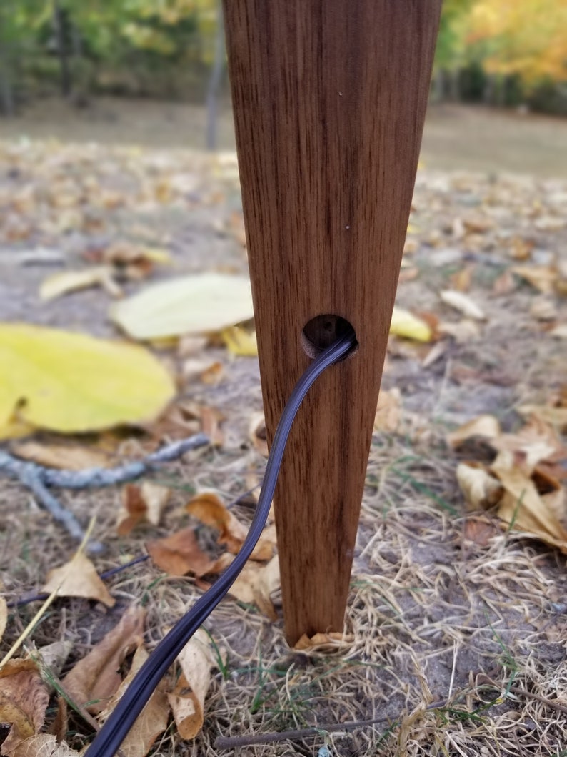 May include: A close-up of a wooden lamp base with a black electrical cord running through a hole in the base. The base is a dark brown color and has a natural wood grain.