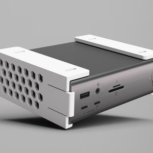 K&ouml;nnte beinhalten: Ein silbernes und schwarzes elektronisches Ger&auml;t mit wei&szlig;en Halterungen. Das Ger&auml;t verf&uuml;gt &uuml;ber mehrere Anschl&uuml;sse und eine wabenf&ouml;rmige Bel&uuml;ftung. Der Markenname "CalDigit" ist sichtbar. Es scheint sich um eine Dockingstation oder ein &auml;hnliches Peripherieger&auml;t zu handeln.