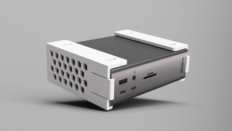 K&ouml;nnte beinhalten: Ein silbernes und schwarzes elektronisches Ger&auml;t mit wei&szlig;en Halterungen. Das Ger&auml;t verf&uuml;gt &uuml;ber mehrere Anschl&uuml;sse und eine wabenf&ouml;rmige Bel&uuml;ftung. Der Markenname "CalDigit" ist sichtbar. Es scheint sich um eine Dockingstation oder ein &auml;hnliches Peripherieger&auml;t zu handeln.