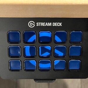 May include: A black Stream Deck with 15 blue buttons, each with a white play button icon. The Stream Deck is mounted on a desk.