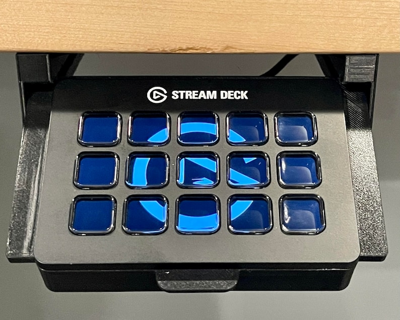 May include: A black Stream Deck with 15 customizable buttons. Each button has a blue, glowing light. The Stream Deck is mounted under a wooden surface.