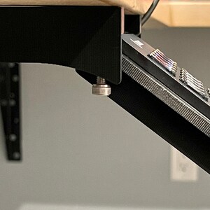 May include: Black desk mount with a silver knob, holding a black device with a row of buttons.