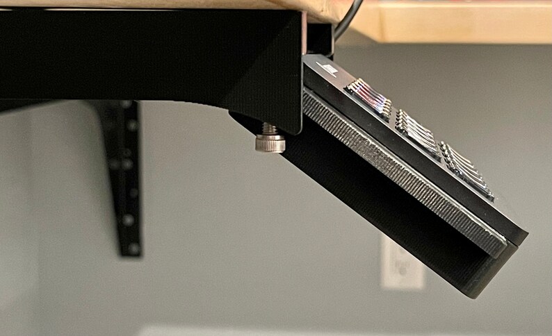 May include: Black desk mount with a silver knob, holding a black device with a row of buttons.