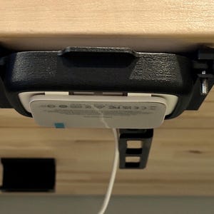 May include: A black plastic mount with a white wireless charger attached to it. The mount is attached to a wooden surface with screws. The charger is plugged in with a white cord.