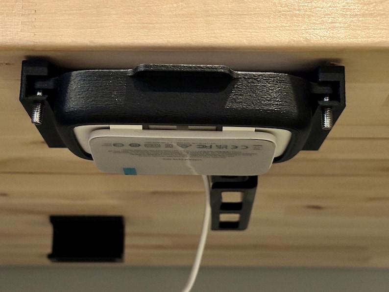 May include: A black plastic mount with a white wireless charger attached to it. The mount is attached to a wooden surface with screws. The charger is plugged in with a white cord.