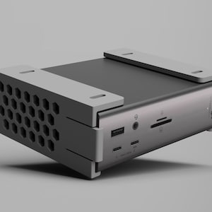 K&ouml;nnte beinhalten: Ein silbernes und graues elektronisches Ger&auml;t mit rechteckiger Form. Das Ger&auml;t hat ein Wabenmuster auf einer Seite und mehrere Anschl&uuml;sse und Steckpl&auml;tze. Hellgraue Akzente befinden sich auf der Oberseite und den Seiten. Der Markenname "CalDigit" ist auf der Seite aufgedruckt.