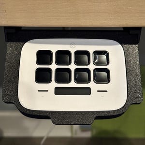 May include: A white, rectangular keypad with a black border and nine black buttons. The keypad has a small, black, circular logo in the top center. The keypad is mounted under a wooden surface.