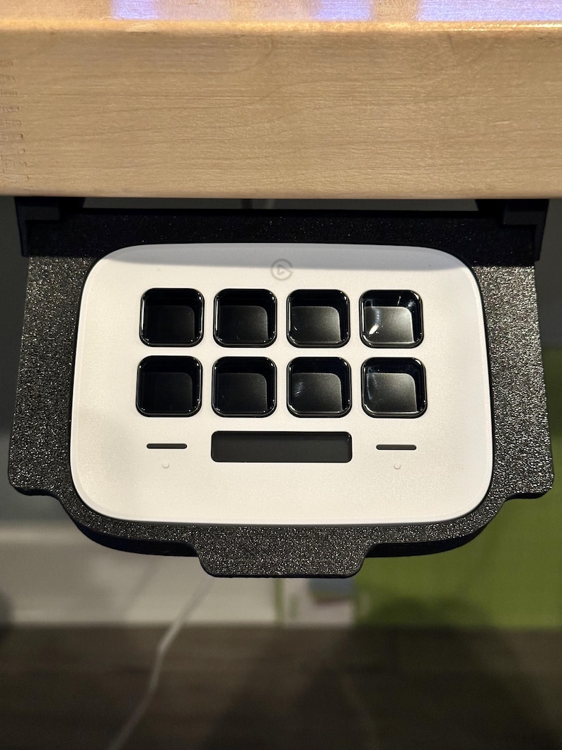 May include: A white, rectangular keypad with a black border and nine black buttons. The keypad has a small, black, circular logo in the top center. The keypad is mounted under a wooden surface.