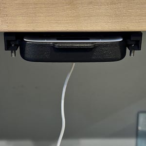May include: A black plastic under-desk drawer with a white interior, mounted to a wooden surface with screws. A white cable hangs from the drawer.