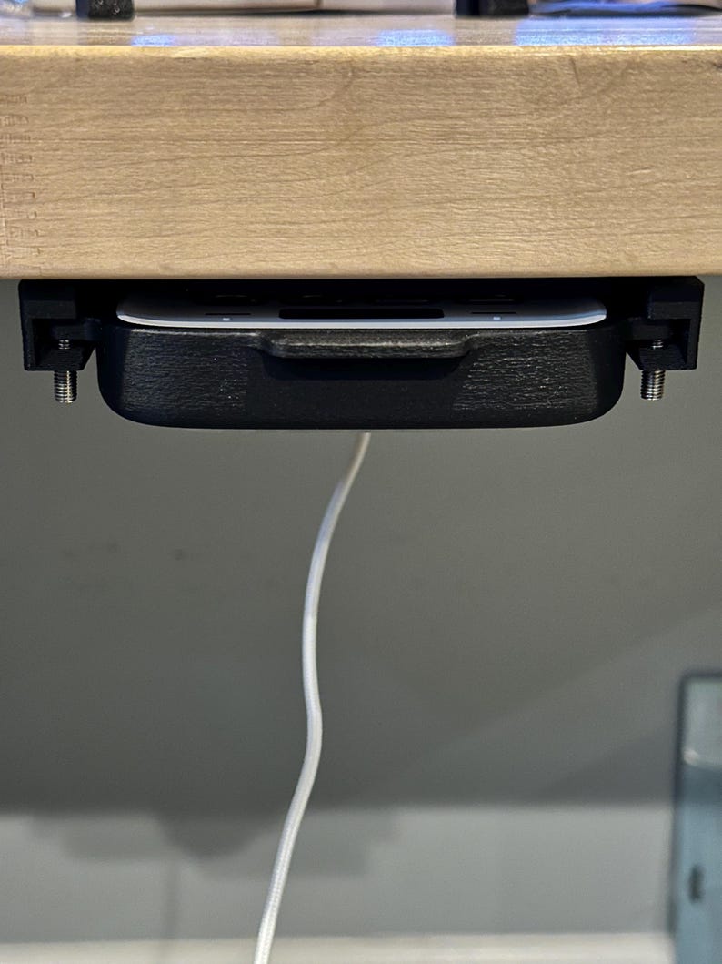 May include: A black plastic under-desk drawer with a white interior, mounted to a wooden surface with screws. A white cable hangs from the drawer.