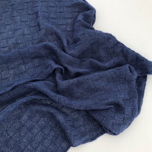 Puede incluir: Una manta tejida azul marino con un patrón texturizado en forma de cuadrícula. La manta está drapeada, mostrando el intrincado tejido y la textura suave. La tela parece ligera y transpirable, adecuada para una variedad de usos.