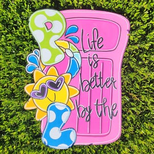 May include: A pink wooden door with the text "Life is better by the pool" and a sun with sunglasses and a blue and white polka dot pool float.