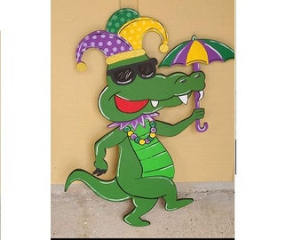May include: A green alligator wearing a purple and gold jester hat and sunglasses, holding a purple and gold umbrella. The alligator is wearing a green and gold beaded necklace.
