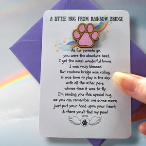 Puede incluir: Una tarjeta blanca con el texto "A Little Hug From Rainbow Bridge" y un dise&ntilde;o de huella de pata. La tarjeta se sostiene en un sobre morado. La tarjeta tiene un poema sobre una mascota.