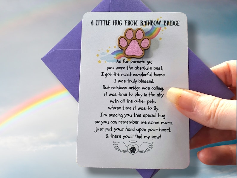 Puede incluir: Una tarjeta blanca con el texto "A Little Hug From Rainbow Bridge" y un dise&ntilde;o de huella de pata. La tarjeta se sostiene en un sobre morado. La tarjeta tiene un poema sobre una mascota.