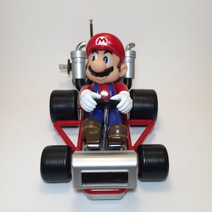 May include: A red and silver toy go-kart with a Mario figure behind the wheel. The go-kart has black wheels and a silver front bumper with a digital clock display.