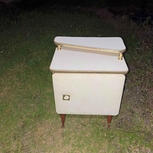 May include: A white bedside table with a tiered top design featuring gold-toned trim along the edges. The unit includes a single cabinet door with a square gold-toned handle and is supported by four tapered brown wooden legs.