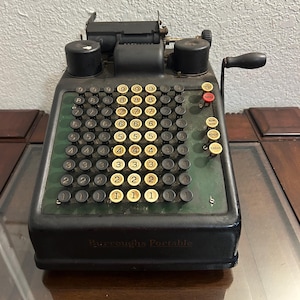 May include: An antique Burroughs Portable adding machine. The machine is black with a green keypad featuring numbered keys and several function buttons. The handle is black, and the text "Burroughs Portable" is visible.