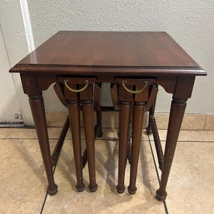 Puede incluir: Un juego de dos mesas nido de madera marrón oscuro. Cada mesa tiene una tapa cuadrada, dos cajones pequeños con tiradores de latón y cuatro patas torneadas. Las mesas están diseñadas para encajar entre sí.