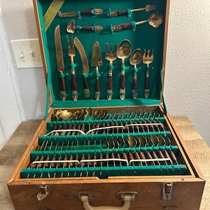 May include: A vintage wooden cutlery case with a green velvet interior. The case is open, revealing a complete set of gold-colored flatware with dark brown wooden handles. The set includes knives, forks, spoons, and serving utensils.