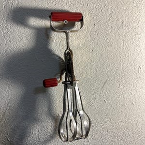 May include: A vintage hand-crank egg beater with red plastic handles and a silver metal whisk. The beater is hanging on a white wall.