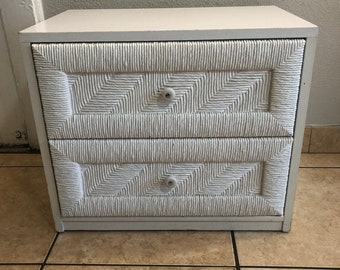 May include: A white two-drawer nightstand with a textured, woven design on the drawer fronts. The nightstand has a simple, rectangular shape with small, round knobs. The overall style is clean and modern.