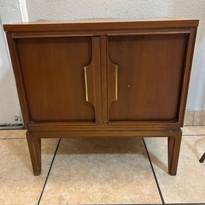 May include: A mid-century modern wooden cabinet with two doors and brass handles. The cabinet has a dark brown finish and sits on four slender legs.
