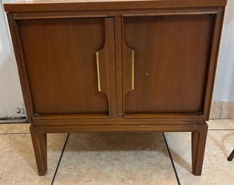 May include: A mid-century modern wooden cabinet with two doors and brass handles. The cabinet has a dark brown finish and sits on four slender legs.