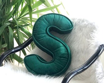 May include: A plush green velvet letter S pillow. The pillow is sitting on a white furry chair.