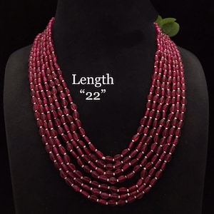 Puede incluir: Un collar de múltiples hilos hecho de cuentas ovaladas de color rojo oscuro. El collar tiene 56 cm de largo. La etiqueta blanca dice "Longitud 22".