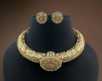 Può includere: Una collana choker dorata con un grande ciondolo rotondo incastonato con pietre bianche. La collana è decorata con motivi floreali e ha un paio di orecchini abbinati.