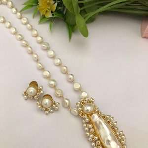 Puede incluir: Un conjunto de collar y pendientes de perlas. El collar presenta una cadena de perlas blancas con una cadena dorada. El colgante tiene una perla grande y alargada rodeada de perlas más pequeñas y detalles dorados. Los pendientes tienen un grupo de perlas.